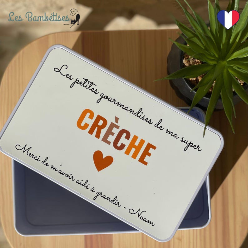 May include: A rectangular, off-white metal box with a lid. The lid features the text "Les petites gourmandises de ma super CR&Egrave;CHE" in black and orange, with a heart symbol. The box is open, revealing a gray interior.