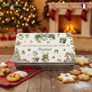 Pode incluir: Uma lata retangular com um design festivo, com um ursinho de pelúcia, árvores de Natal e o texto "Les petites douceurs de Noël de Raphaël". A lata é rodeada por vários biscoitos de Natal numa superfície de madeira.