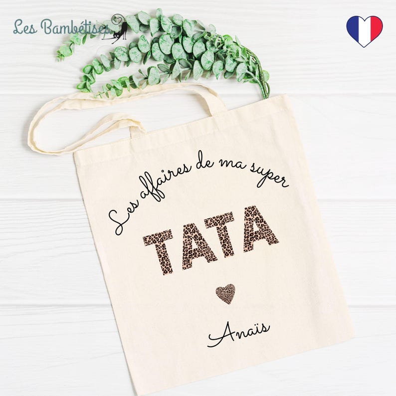 Op de afbeelding: Een witte tote bag met zwarte tekst die "Les affaires de ma super TATA" zegt met een luipaardprint. De tekst "Anais" staat in cursief onder de luipaardprint.