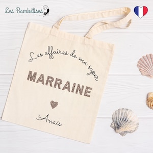Puede incluir: Una bolsa de tela blanca con texto negro que dice "Les affaires de ma super MARRAINE Anais". El texto está en una fuente cursiva y hay un pequeño corazón con un diseño de estampado de leopardo debajo del texto.