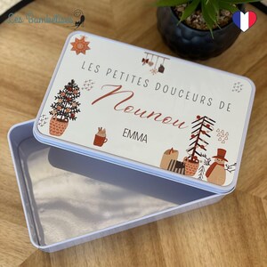Könnte beinhalten: Eine rechteckige, offene, hellblaue Blechdose mit dekorativem Deckel. Der Deckel zeigt den Text "LES PETITES DOUCEURS DE Nounou EMMA" in einer Schreibschrift, zusammen mit festlichen Illustrationen von Weihnachtsbaum, Schneemann und anderen Elementen.