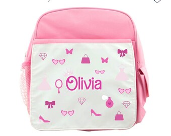 Könnte beinhalten: Rosa Rucksack mit einer weißen Fronttasche, die mit einem rosa-weißen Design aus Krone, Spiegel, Kleid, Handtasche, Sonnenbrille, Schmetterlingen und Diamanten verziert ist. Der Name Olivia ist in rosa Buchstaben auf der Fronttasche geschrieben.