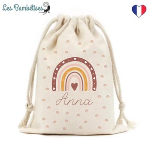 Puede incluir: Una bolsa de cordón beige con un diseño de arcoíris y el nombre "Anna" impreso en ella. El arcoíris está formado por rayas rosas, naranjas y amarillas con contornos marrones y corazones. La bolsa está cubierta con un patrón de pequeños corazones rosas.