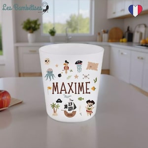 Puede incluir: Vaso blanco decorado con ilustraciones de temática pirata, incluyendo un barco pirata, un mapa del tesoro y varias criaturas marinas. El nombre "MAXIME" está impreso en marrón. El vaso tiene una forma ligeramente acampanada.