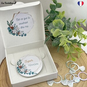 Peut inclure: Une boîte cadeau blanche ouverte, contenant un badge rond avec l'inscription dorée "Super Témoin". Le badge est orné d'un motif floral avec de l'eucalyptus et du coton. À l'intérieur, un message en français : "Est-ce que tu voudrais être ma..."