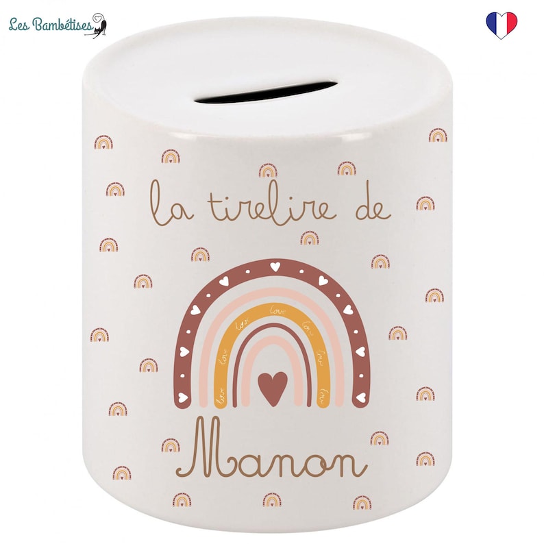 May include: White ceramic money box with a rainbow design and the text "la tirelire de Manon" in brown lettering. The rainbow is made up of pink, orange, and yellow stripes with brown hearts around it. The background is white with a brown polka dot pattern.