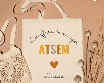 Puede incluir: Una bolsa de tela blanca con texto negro que dice "Les affaires de ma super ATSEM Louison". El texto está en una fuente de escritura a mano. La bolsa tiene un corazón marrón en el centro. La bolsa está sobre una superficie marrón claro.