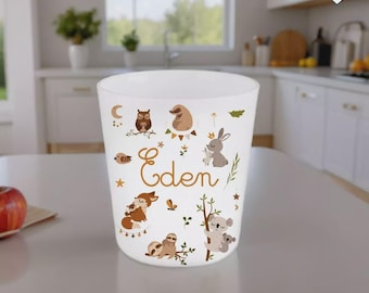 Può includere: Un bicchiere bianco decorato con illustrazioni di animali del bosco e il nome "Eden" in oro. Il design include un gufo marrone, un coniglio, un koala e un bradipo. Il bicchiere è su una superficie bianca.