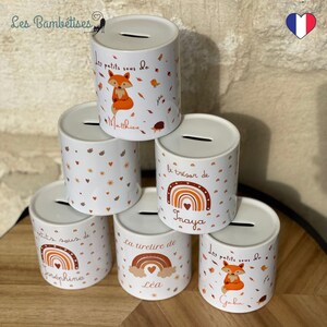 May include: Five white ceramic money boxes with different designs. The designs include a fox, a rainbow, and hearts. Each money box has a name written on it: Matthieu, Inaya, Josephine, Lea, and Gabin.