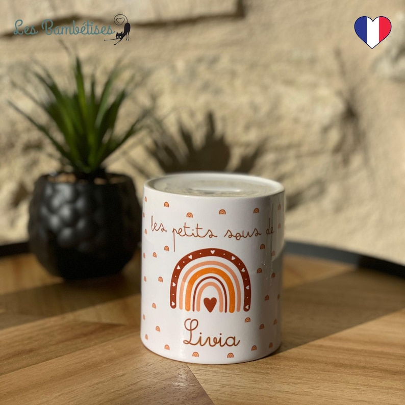 May include: White ceramic money box with a rainbow design and the text "les petits sous de Livia".
