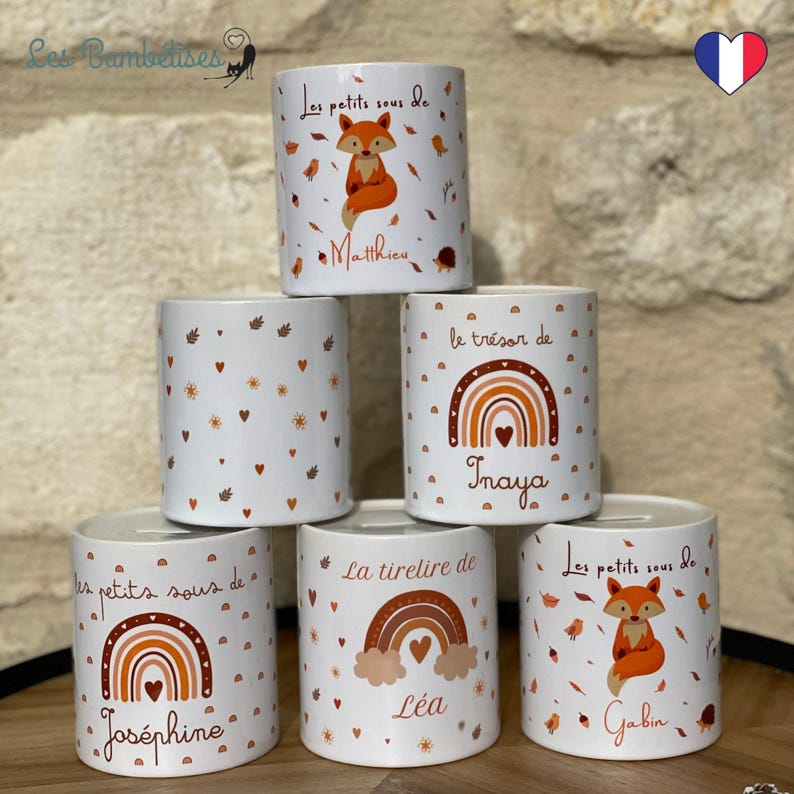 May include: Six white ceramic money boxes with different designs. The designs include a fox, a rainbow, and hearts. Each money box has a name on it, including Inaya, L&eacute;a, Jos&eacute;phine, Gabin, and Matthieu.