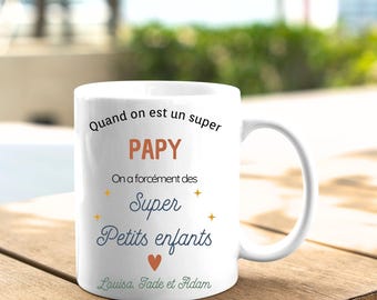 May include: White ceramic mug with a blue and orange design. The mug says "Quand on est un super PAPY On a forcément des Super Petits enfants Louisa, Tade et Adam".