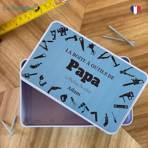 Puede incluir: Una caja de herramientas de metal azul y blanco con el texto "LA BOITE À OUTILS DE Papa Le Meilleur Bricoleur Adam" en la tapa. La tapa está decorada con un círculo de siluetas negras de herramientas.