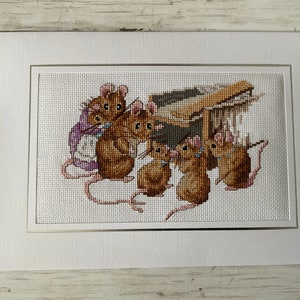 Peut inclure: Une broderie au point de croix représentant un groupe de souris brunes avec des nœuds bleus, debout devant une structure en bois. Les souris regardent le spectateur.