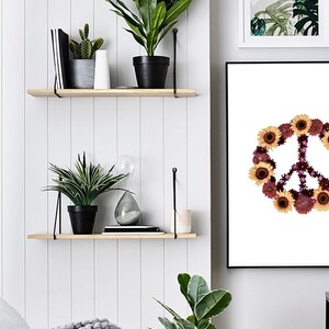May include: A white framed print with a peace sign made of flowers. The peace sign is surrounded by sunflowers and other flowers in shades of purple, pink, and yellow. The print is hanging on a white wall with two wooden shelves above it. The shelves are decorated with plants and other items.