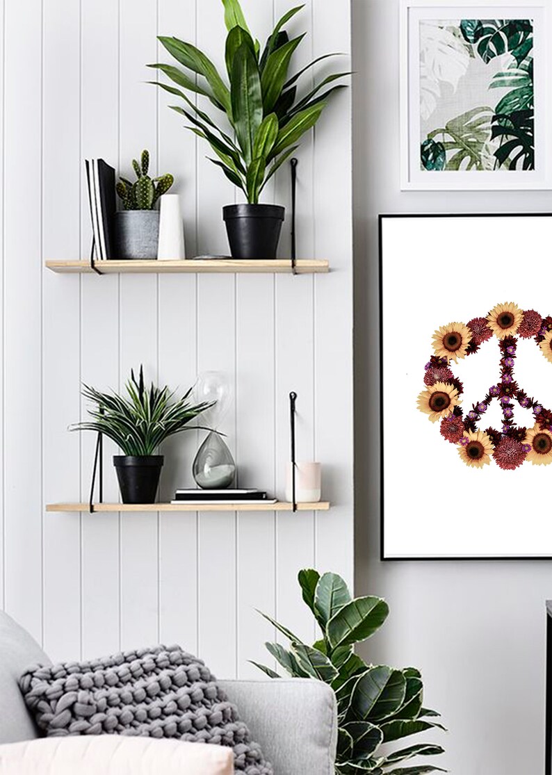 May include: A white framed print with a peace sign made of flowers. The peace sign is surrounded by sunflowers and other flowers in shades of purple, pink, and yellow. The print is hanging on a white wall with two wooden shelves above it. The shelves are decorated with plants and other items.