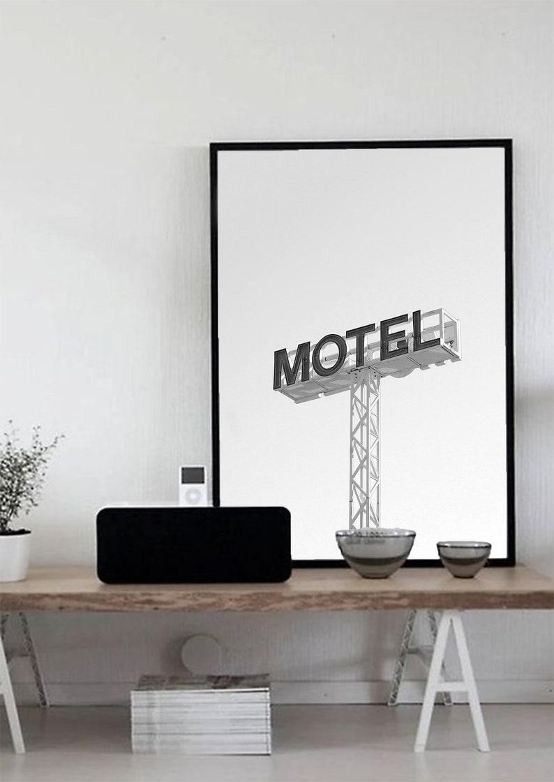 May include: A black and white photograph of a motel sign with the word "MOTEL" in bold letters. The sign is mounted on a tall metal pole. The photograph is framed in a black frame and is hanging on a white wall above a wooden table.