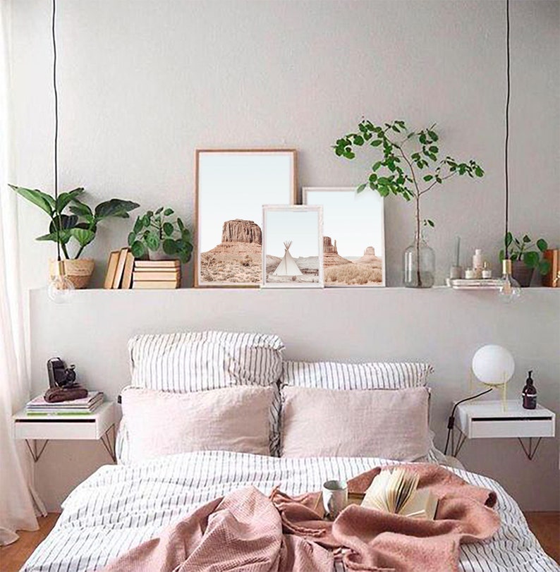 May include: A bedroom with a white bed with a pink blanket and striped bedding. There are three framed prints above the bed with desert landscapes. The room has a white shelf with plants and a nightstand on each side of the bed.