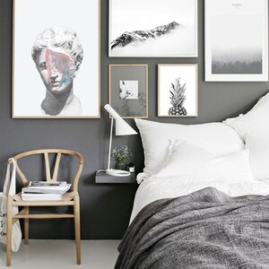 May include: A bedroom interior with a white bed, a wooden chair, and a gray wall with black and white framed artwork. The artwork includes a bust of a person, a mountain range, and a pineapple.