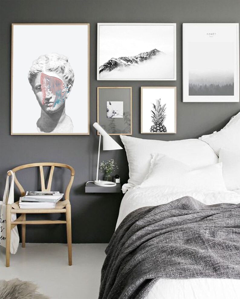 May include: A bedroom interior with a white bed, a wooden chair, and a gray wall with black and white framed artwork. The artwork includes a bust of a person, a mountain range, and a pineapple.