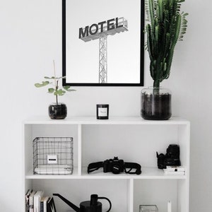 May include: A black and white photograph of a motel sign with the word "MOTEL" in bold letters. The photograph is framed in a black frame and is hanging on a white wall above a white shelf with various items, including a potted plant, a watering can, and a camera.