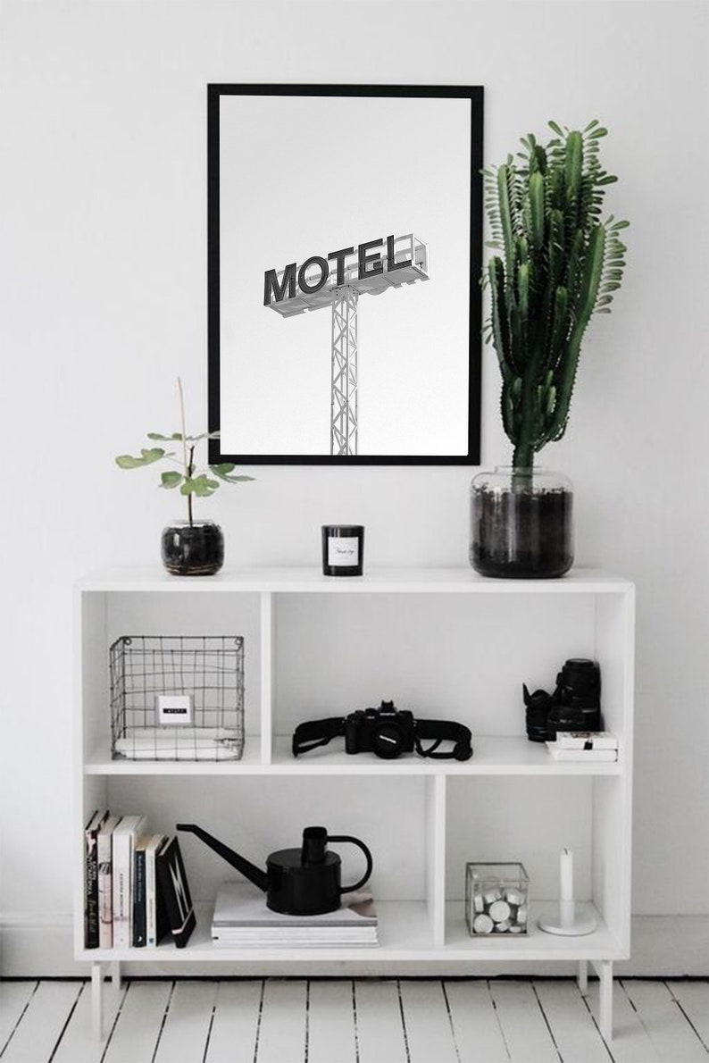 May include: A black and white photograph of a motel sign with the word "MOTEL" in bold letters. The photograph is framed in a black frame and is hanging on a white wall above a white shelf with various items, including a potted plant, a watering can, and a camera.