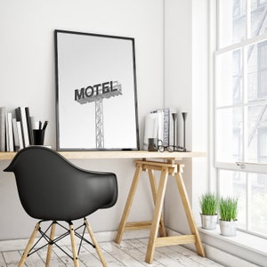 May include: A black and white photograph of a motel sign with the word "MOTEL" in bold letters. The photo is framed and hanging on a white wall above a wooden desk with a black chair and a wooden easel.