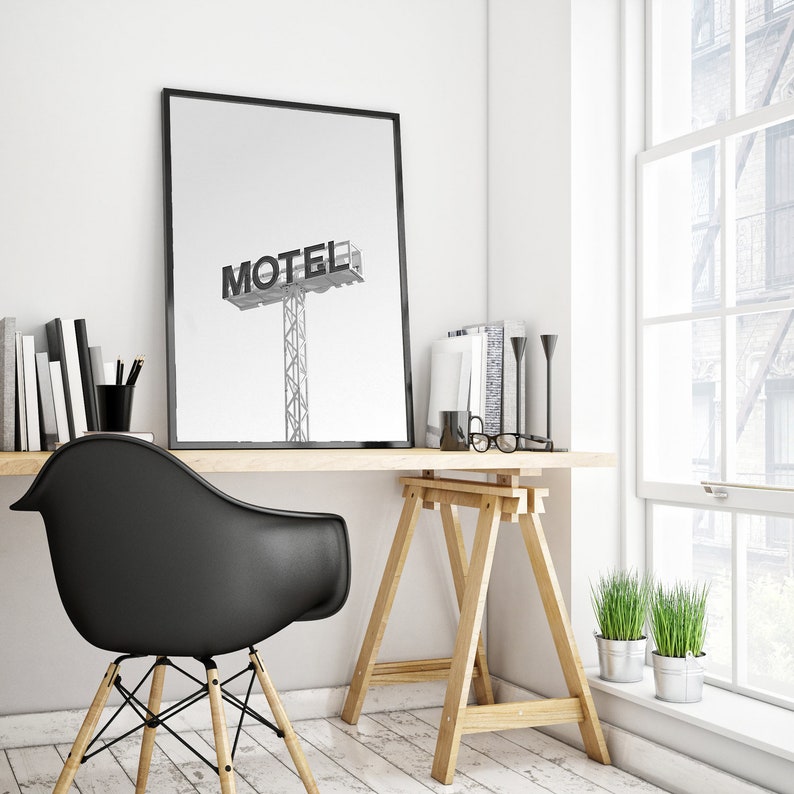 May include: A black and white photograph of a motel sign with the word "MOTEL" in bold letters. The photo is framed and hanging on a white wall above a wooden desk with a black chair and a wooden easel.