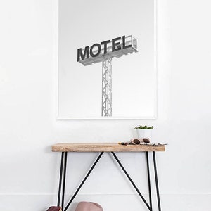 May include: A black and white photograph of a motel sign with the word "MOTEL" in bold letters. The sign is mounted on a tall metal pole. The photo is framed in a white frame and hanging on a white wall above a wooden console table with black metal legs.