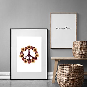 May include: A framed print of a peace sign made of dried flowers, including sunflowers and purple flowers. The print is hanging on a gray wall next to a framed print with the word "breathe" written in black ink.
