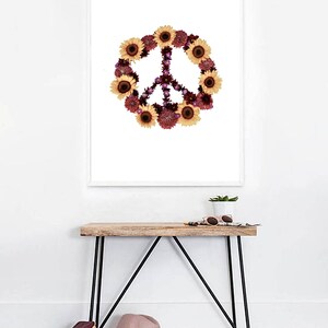 May include: A white framed print with a peace sign made of sunflowers and other flowers on a white background. The print is hanging on a white wall above a wooden table with black metal legs. There is a potted plant, sunglasses, and a book on the table.