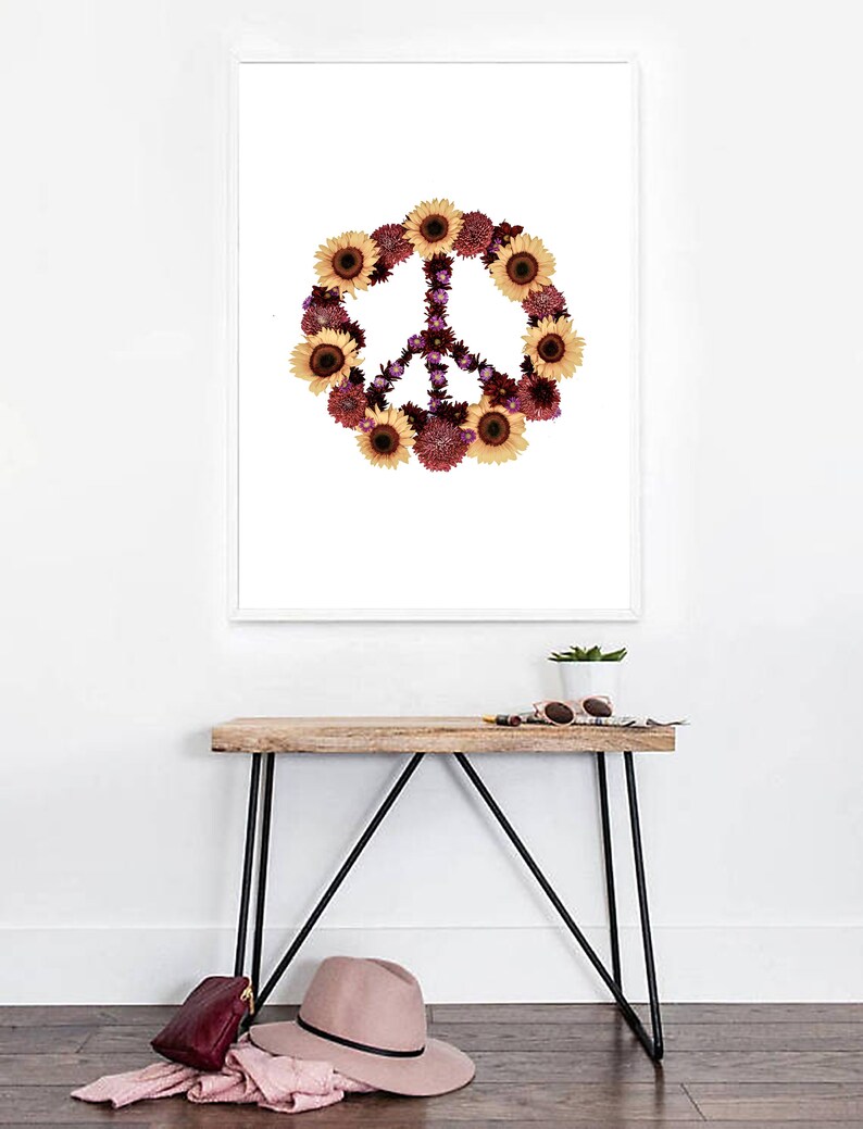 May include: A white framed print with a peace sign made of sunflowers and other flowers on a white background. The print is hanging on a white wall above a wooden table with black metal legs. There is a potted plant, sunglasses, and a book on the table.