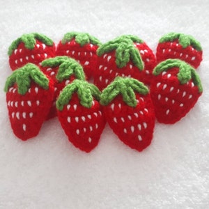 Puede incluir: Un grupo de siete fresas de ganchillo en tonos rojos y verdes. Las fresas están dispuestas en un racimo sobre una superficie blanca.