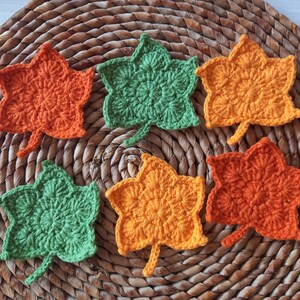 May include: Six handmade crochet leaf coasters in shades of orange and green. The coasters are arranged on a woven, circular placemat. Each leaf has a detailed, textured pattern and a small stem.