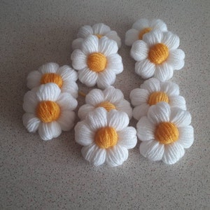Puede incluir: Un grupo de 10 flores de margarita blancas de ganchillo con centros amarillos. Las flores están dispuestas en un grupo sobre una superficie gris claro.