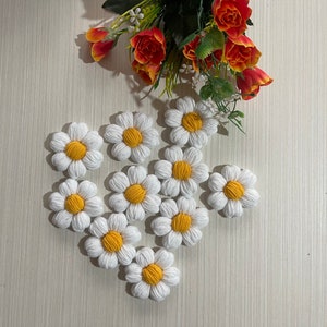 Op de afbeelding: Een verzameling van tien witte gehaakte madeliefjes met gele centra. De bloemen zijn in de vorm van een hart op een licht houten oppervlak gerangschikt.