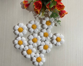 May include: A collection of ten white crocheted daisy flowers with yellow centers. The flowers are arranged in a heart shape on a light wood surface.