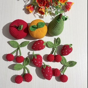 May include: A collection of crocheted fruit, including a red apple, an orange, a green pear, strawberries, and cherries. The fruit is arranged on a white surface.