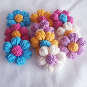 May include: A collection of nine crocheted flower embellishments in various colors, including pink, blue, yellow, and white. The flowers are made with yarn and have a three-dimensional appearance.