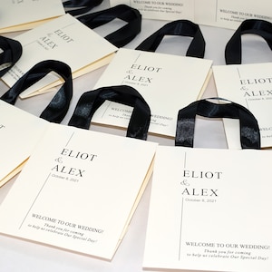 K&ouml;nnte beinhalten: Wei&szlig;e Geschenktaschen mit schwarzen Schleifenb&auml;ndern und einem bedruckten Design, auf dem "Eliot & Alex" und "8. Oktober 2021" steht. Die Taschen haben auch den Text "Willkommen zu unserer Hochzeit! Vielen Dank, dass Sie gekommen sind, um uns bei der Feier unseres besonderen Tages zu helfen!"