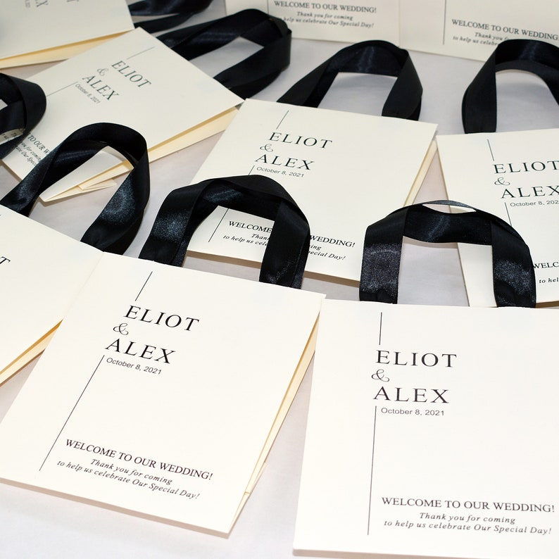 K&ouml;nnte beinhalten: Wei&szlig;e Geschenktaschen mit schwarzen Schleifenb&auml;ndern und einem bedruckten Design, auf dem "Eliot & Alex" und "8. Oktober 2021" steht. Die Taschen haben auch den Text "Willkommen zu unserer Hochzeit! Vielen Dank, dass Sie gekommen sind, um uns bei der Feier unseres besonderen Tages zu helfen!"