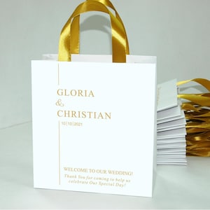 K&ouml;nnte beinhalten: Wei&szlig;e Papier-Geschenk-Tasche mit goldenen Schleifenb&auml;ndern. Die Tasche hat goldene Schrift, die "GLORIA & CHRISTIAN 10/10/2021" und "WELCOME TO OUR WEDDING! Thank You for coming to help us celebrate Our Special Day!" lautet.