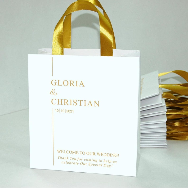 K&ouml;nnte beinhalten: Wei&szlig;e Papier-Geschenk-Tasche mit goldenen Schleifenb&auml;ndern. Die Tasche hat goldene Schrift, die "GLORIA & CHRISTIAN 10/10/2021" und "WELCOME TO OUR WEDDING! Thank You for coming to help us celebrate Our Special Day!" lautet.