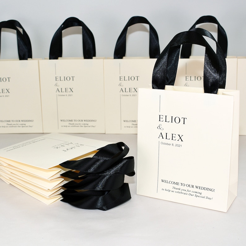 K&ouml;nnte beinhalten: Ein Stapel elfenbeinfarbener Hochzeitstaschen mit schwarzen Satinb&auml;ndern als Henkel. Die Taschen sind mit dem Text "Eliot & Alex" und dem Datum "8. Oktober 2021" bedruckt. Die Taschen haben ein schwarz-wei&szlig;es Grafikdesign mit einer vertikalen Linie und dem Text "Willkommen zu unserer Hochzeit! Vielen Dank, dass Sie gekommen sind, um uns bei der Feier unseres besonderen Tages zu helfen!"