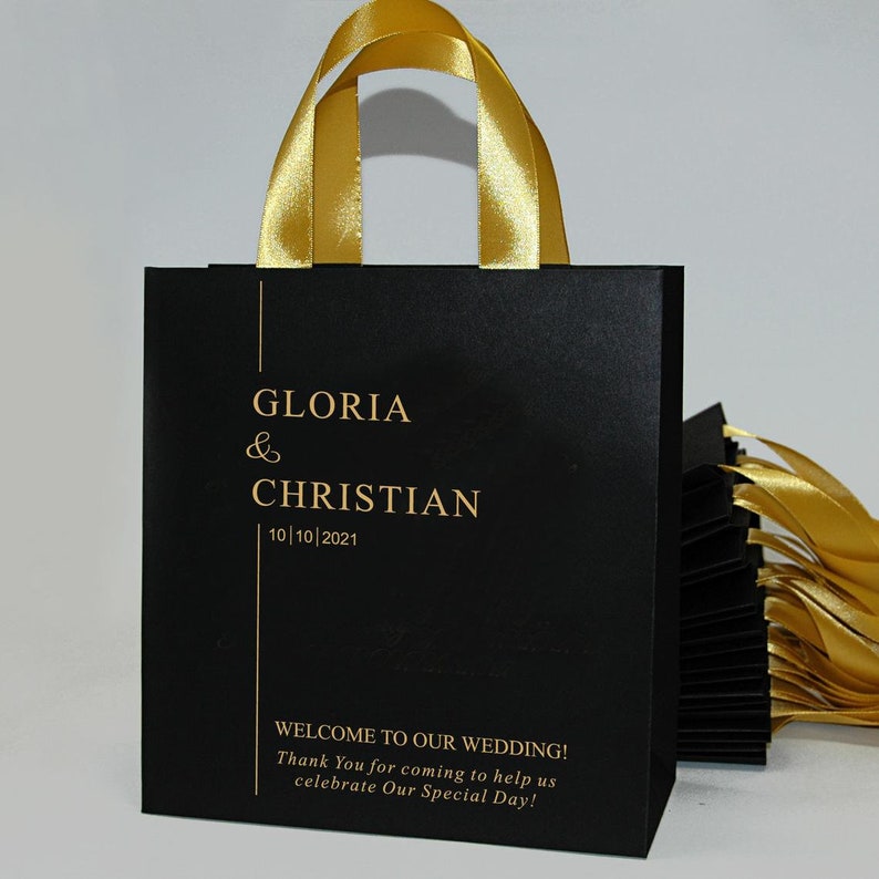 K&ouml;nnte beinhalten: Schwarze Geschenkt&uuml;te mit goldenen Schleifenb&auml;ndern als Henkel. Die Tasche tr&auml;gt den Text "GLORIA & CHRISTIAN 10/10/2021" und "WELCOME TO OUR WEDDING! Thank You for coming to help us celebrate Our Special Day!" in Gold.