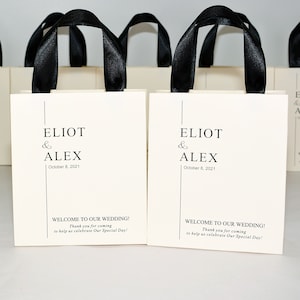 K&ouml;nnte beinhalten: Wei&szlig;e Geschenktaschen mit schwarzen Schleifenb&auml;ndern und dem Text "ELIOT & ALEX October 8, 2021" und "WELCOME TO OUR WEDDING! Thank you for coming to help us celebrate Our Special Day!"