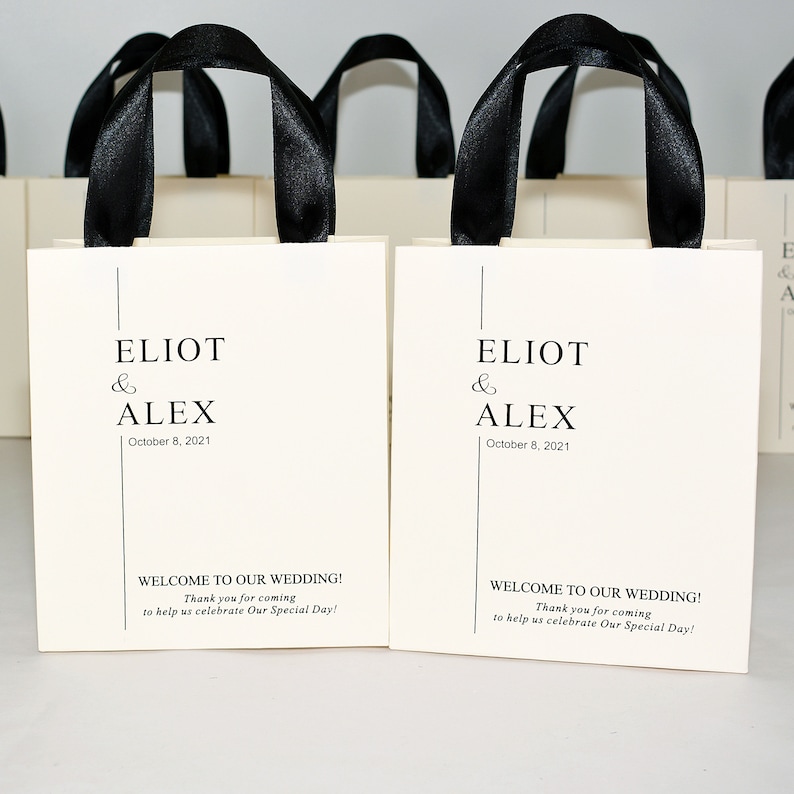 K&ouml;nnte beinhalten: Wei&szlig;e Geschenktaschen mit schwarzen Schleifenb&auml;ndern und dem Text "ELIOT & ALEX October 8, 2021" und "WELCOME TO OUR WEDDING! Thank you for coming to help us celebrate Our Special Day!"