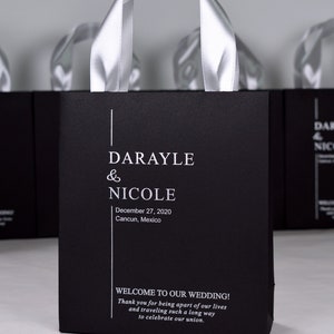 K&ouml;nnte beinhalten: Schwarze und wei&szlig;e Hochzeitstasche mit einem silbernen Schleifenhenkel. Die Tasche tr&auml;gt die Namen "Darayle & Nicole" und das Datum "27. Dezember 2020". Die Tasche tr&auml;gt auch den Text "Willkommen zu unserer Hochzeit! Vielen Dank, dass Sie Teil unseres Lebens sind und so weit gereist sind, um unsere Vereinigung zu feiern."