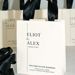 K&ouml;nnte beinhalten: Wei&szlig;e Geschenktaschen mit schwarzen Schleifenb&auml;ndern als Henkel. Die Taschen haben den Text "ELIOT & ALEX October 8, 2021" auf der Vorderseite gedruckt. Die Taschen haben auch den Text "WELCOME TO OUR WEDDING! Thank you for coming to help us celebrate Our Special Day!" auf der Vorderseite gedruckt.