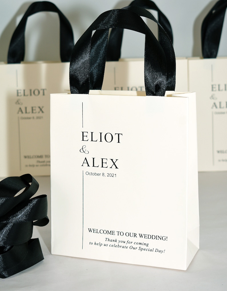K&ouml;nnte beinhalten: Wei&szlig;e Geschenktaschen mit schwarzen Schleifenb&auml;ndern als Henkel. Die Taschen haben den Text "ELIOT & ALEX October 8, 2021" auf der Vorderseite gedruckt. Die Taschen haben auch den Text "WELCOME TO OUR WEDDING! Thank you for coming to help us celebrate Our Special Day!" auf der Vorderseite gedruckt.